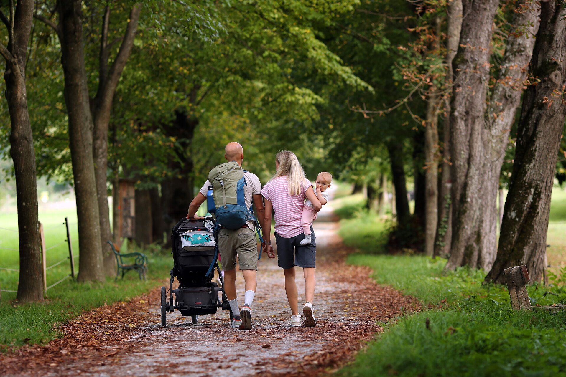 Eine Familie spaziert mit einem Kinderwagen einen Schotterweg entlang. 