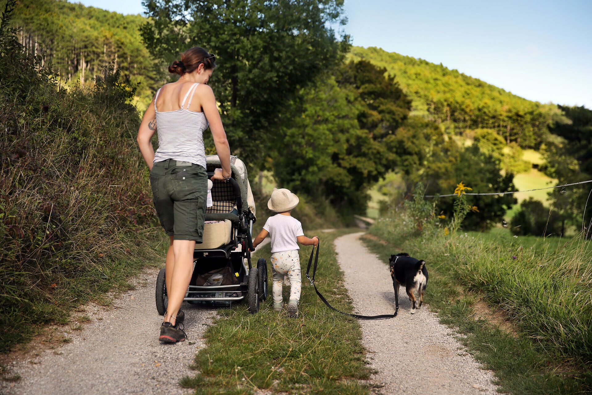 Eine Frau schiebt einen Kinderwagen, ihr Kleinkind hält einen Hund an der Leine und geht neben ihr her. 