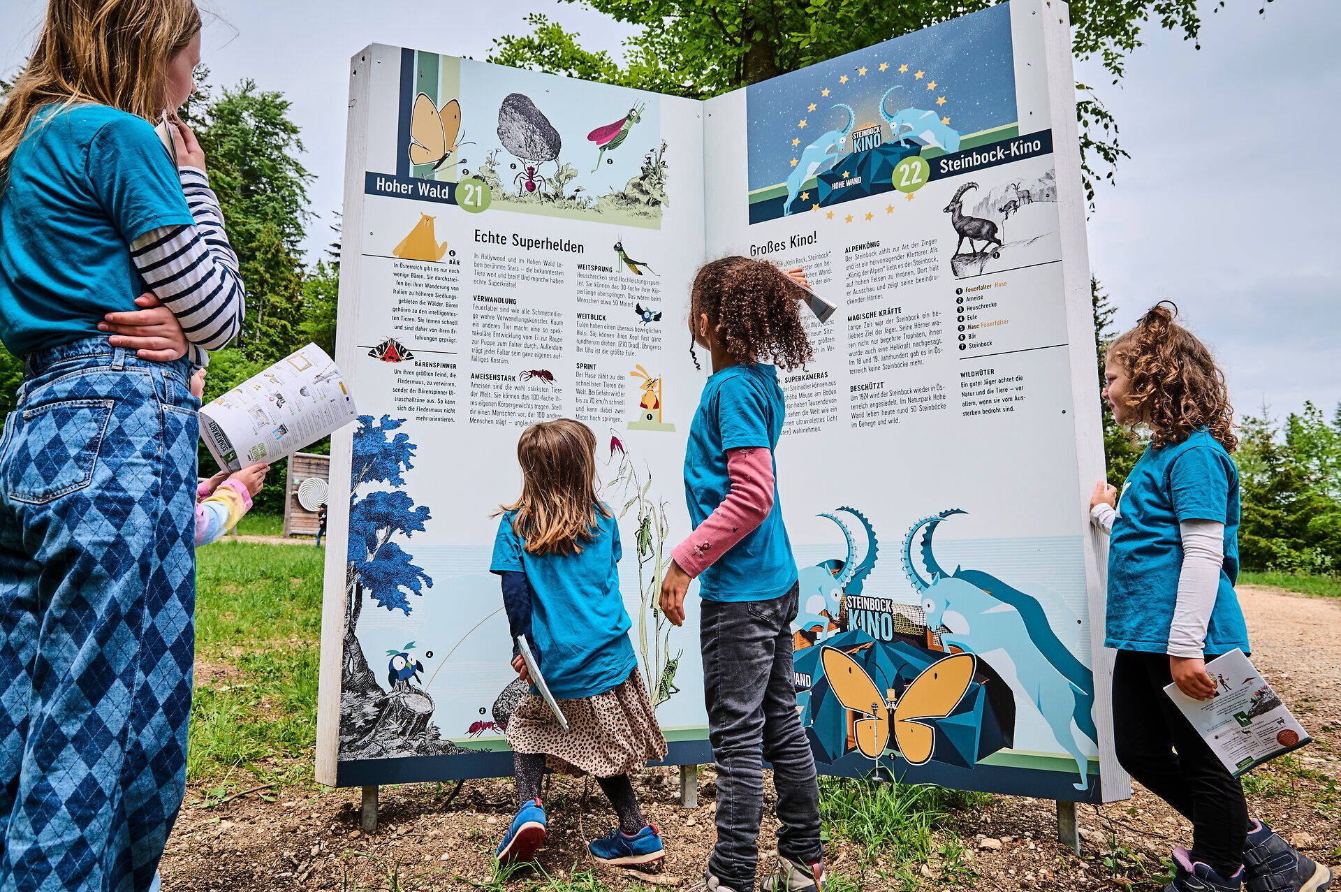Eine Gruppe von Kindern erkundet neugierig die interaktiven Stationen, die spannende Informationen über die Natur und die Umgebung bieten. Die fröhlichen Gesichter und die bunten T-Shirts spiegeln die Freude am Lernen und Entdecken wider, während die sanfte Brise durch die Bäume weht und die Schönheit der Landschaft um sie herum verstärkt.