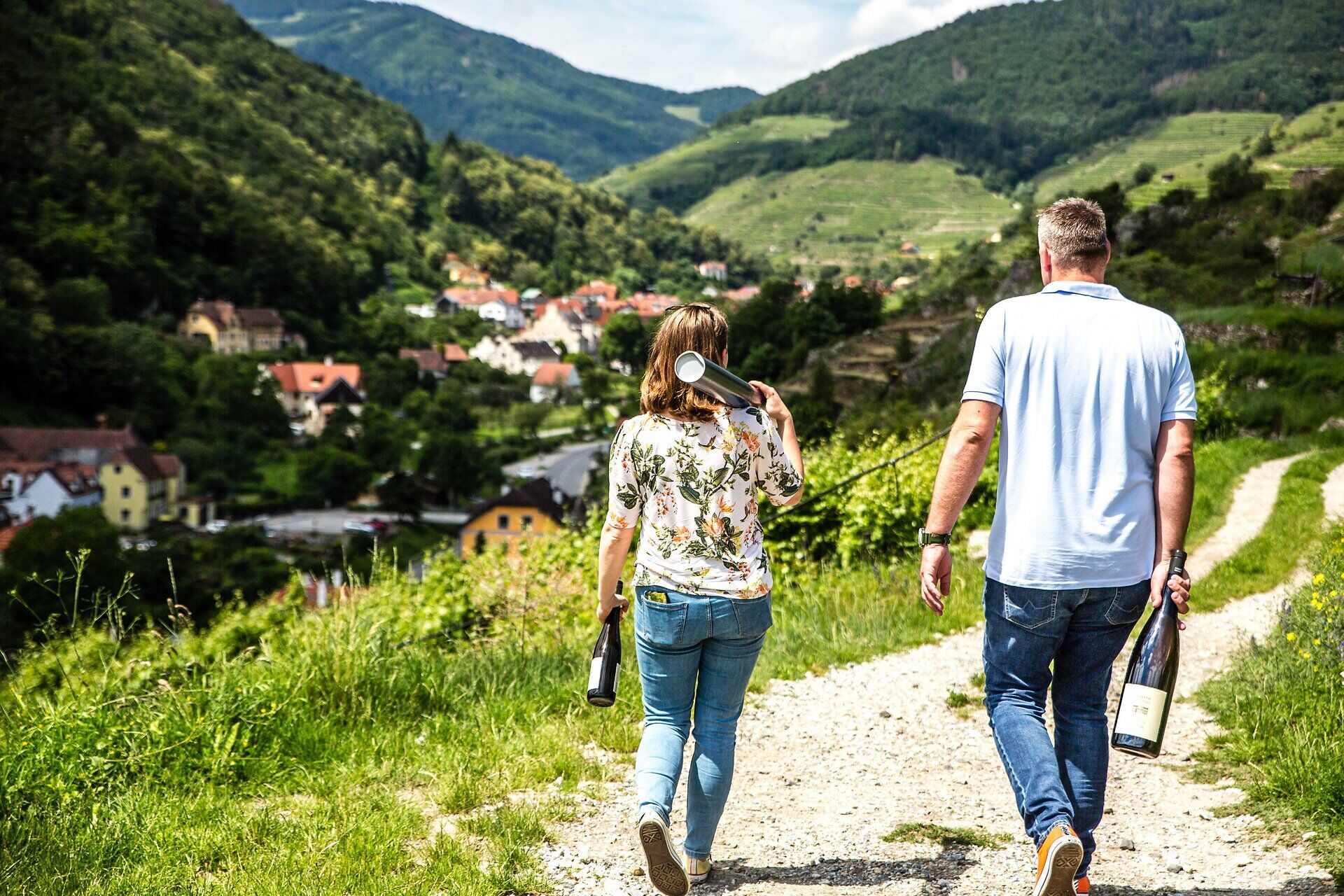 Ein entspannter Spaziergang durch die malerische Landschaft, umgeben von sanften Hügeln und Weinreben, lädt dazu ein, die Schönheit des Sommers zu genießen. Die frische Luft und der Duft der Natur schaffen eine harmonische Atmosphäre, während die beiden Wanderer mit einem Lächeln die Aussicht auf die charmante Ortschaft genießen.