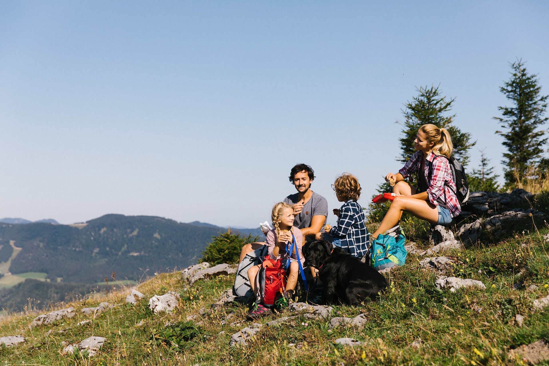 Eine fröhliche Familie genießt die atemberaubende Aussicht auf die umliegenden Berge, während sie eine wohlverdiente Pause auf einem Felsen einlegt. Der Hund liegt entspannt zu ihren Füßen und die Kinder lachen, während sie die Natur erkunden. Diese harmonische Szene verkörpert die Freude am Wandern und die Schönheit des Mostviertels.
