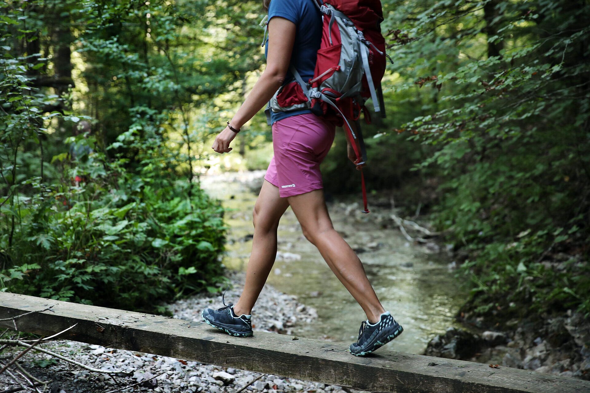 Frau in Wanderoutfit balanciert über Holzpfosten über einen kleinen Bach. 