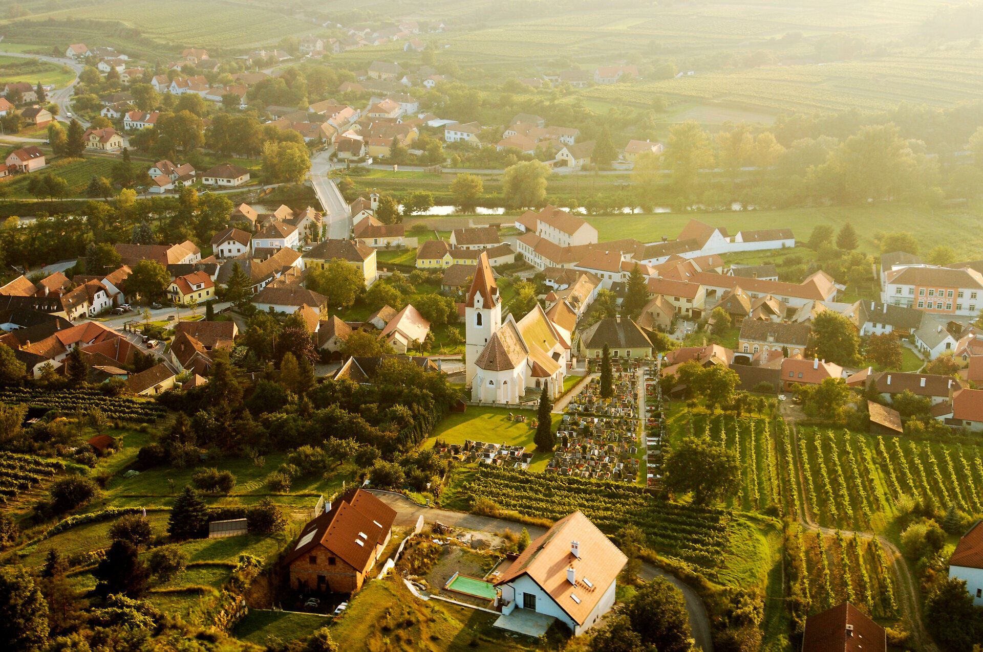 Sanfte Hügel und grüne Weinreben umarmen das malerische Dorf, während die warmen Sonnenstrahlen die Landschaft in goldenes Licht tauchen. Die idyllische Atmosphäre lädt dazu ein, die Ruhe der Natur zu genießen und die Schönheit der Umgebung zu erkunden.