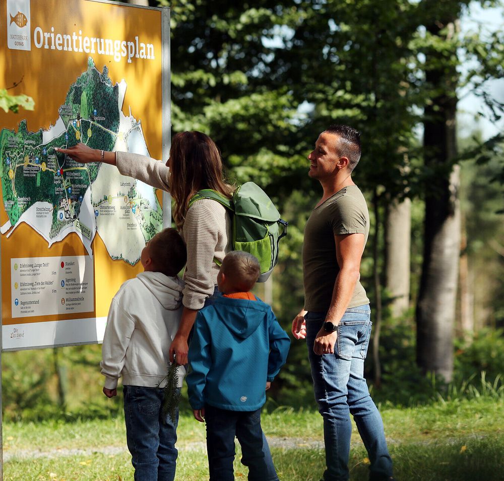 Eine Familie steht vor dem Orientierungsplan im Naturpark Geras, welcher auf einer großen Tafel abgebildet ist. 