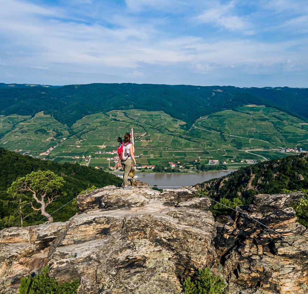 Ein Wanderer steht auf einem majestätischen Felsen und genießt den atemberaubenden Blick über die sanften Hügel und das glitzernde Wasser des Flusses. Die üppige grüne Landschaft erstreckt sich bis zum Horizont und vermittelt ein Gefühl von Freiheit und Abenteuer. Hier, in der Natur, wird der Sommer in seiner schönsten Form erlebbar.